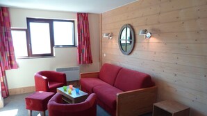 Una habitación con un sofá rojo, un sillón rojo, una mesita con tazas de colores, un espejo en la pared y una ventana con cortinas.