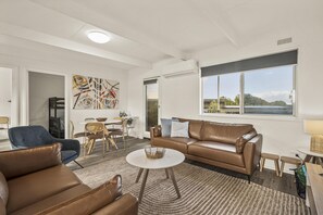 A modern living room with a brown leather sofa, a blue armchair, and a dining area with a table and chairs. - The HardyOne - Aldinga Beach - C21 SouthCoast Holidays (Aldinga Beach)