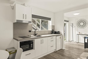 A modern kitchen with white cabinetry, a built-in oven, a stainless steel countertop, and a window with a view of greenery. - The HardyOne - Aldinga Beach - C21 SouthCoast Holidays (Aldinga Beach)