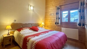 Una camera da letto con soffitto in legno, un letto con copripiumino rosso e bianco, due lampade da comodino, una finestra con tende e vista sugli edifici esterni.