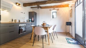 A modern kitchen with wooden beams, a dining area with white chairs, and a small TV on a stand. - Charming Renovated House with Garden in the Heart of Salt Marshes, Close to Guérande and La Baule (Guérande)