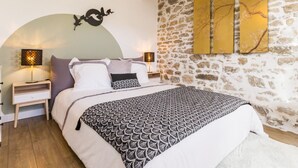 A bedroom with a stone wall, a bed with patterned bedding, two bedside lamps, and wall art. - Charming Renovated House with Garden in the Heart of Salt Marshes, Close to Guérande and La Baule (Guérande)