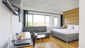 A modern hotel room with a sofa, a bed, a small table with a remote control, and a window with a view of the outside.