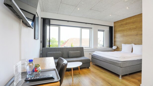 Ein modernes Hotelzimmer mit einer Couch, einem Bett, einem kleinen Tisch mit Fernbedienung und einem Fenster mit Blick ins Freie.