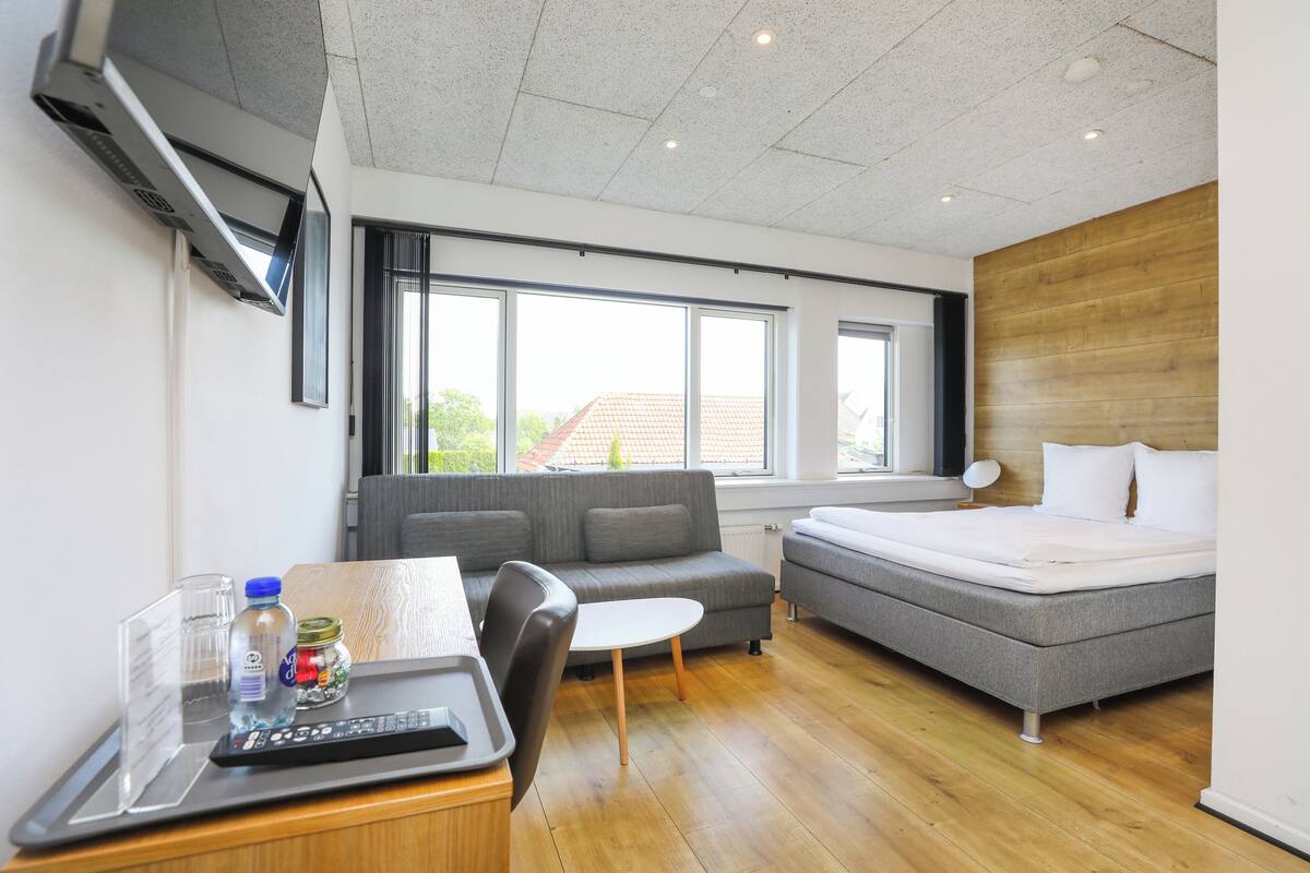 A modern hotel room with a sofa, a bed, a small table with a remote control, and a window with a view of the outside.