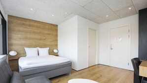 Ein modernes Hotelzimmer mit einem hölzernen Kopfteil, einem grauen Sofa, einem weißen Bett, einem kleinen Tisch und einer Tür mit einem Schild.