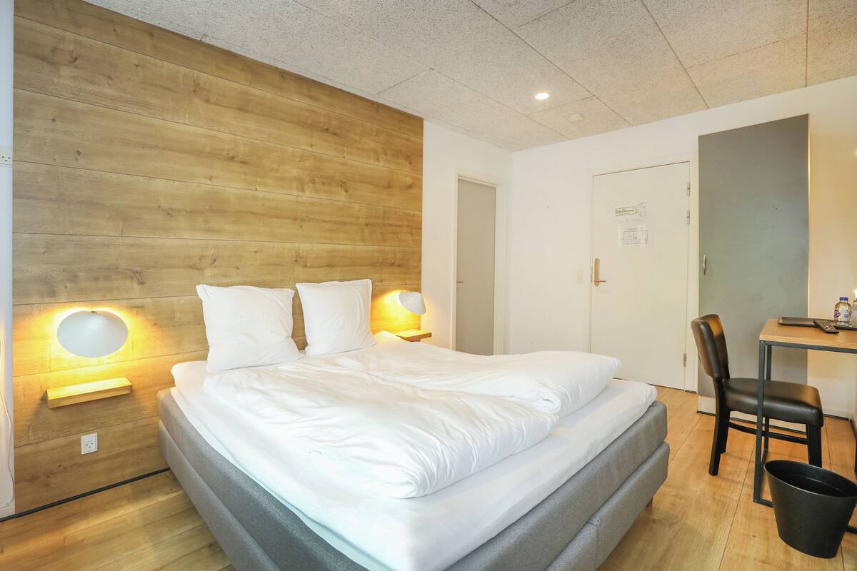 A modern hotel room with a wooden accent wall, a neatly made bed, a desk with a chair, and a small trash bin.