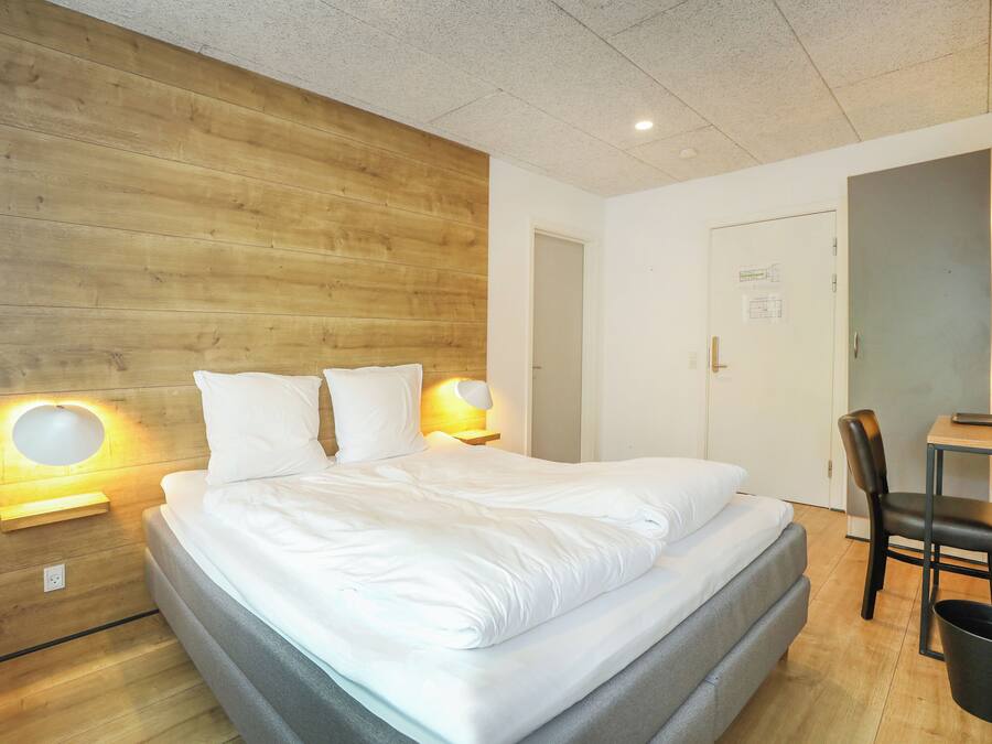 Ein modernes Hotelzimmer mit einer Wand in Holzoptik, einem ordentlich bezogenen Bett, einem Schreibtisch mit Stuhl und einer kleinen Mülltonne.
