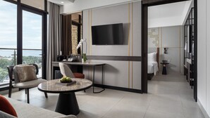 A modern hotel room with a large window, a flat-screen TV, a dining area with a round table, and a separate bedroom with a bed and a nightstand. - Hotel Santika Premiere Padang (Padang)
