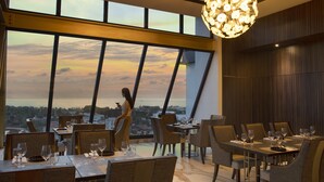 A restaurant with large windows offering a view of the cityscape at sunset. - Hotel Santika Premiere Padang (Padang)