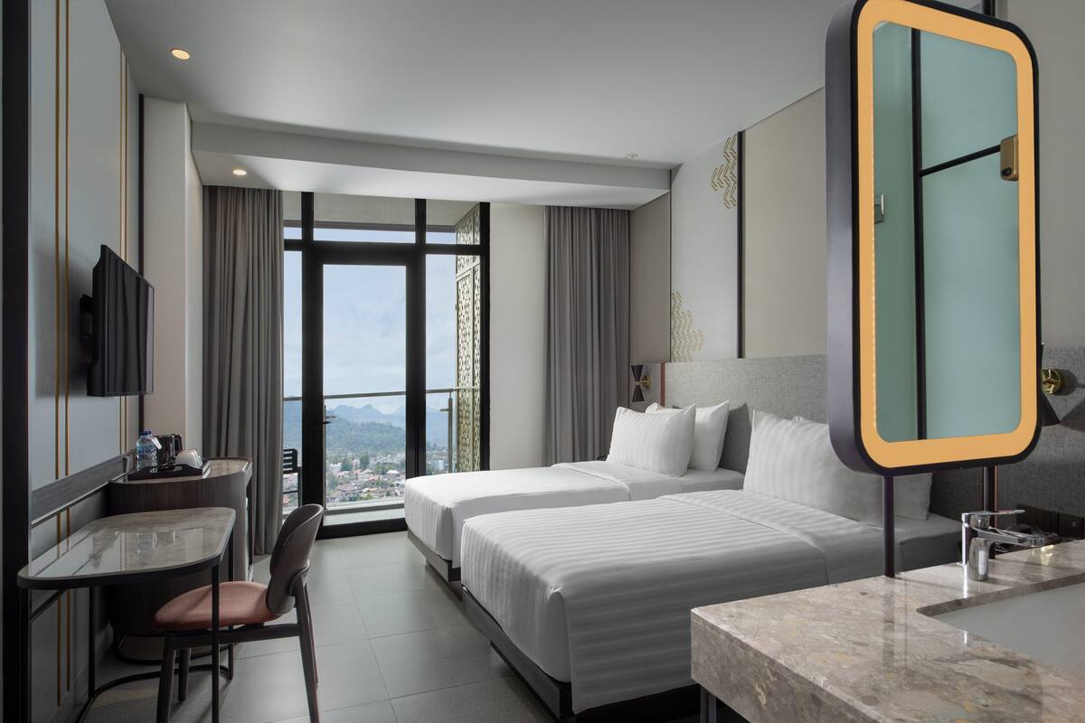 Habitación de hotel con dos camas, un escritorio con silla, un espejo grande y vistas a la ciudad a través de amplias ventanas.