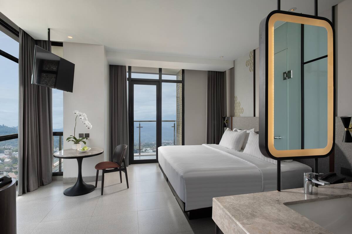 Una habitación de hotel moderna con una cama grande, una mesa redonda con una silla, un televisor montado en la pared y un espejo grande con un marco original.