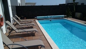 Et rektangulært pool med klart, blåt vand, omgivet af en fliseterrasse. Der er flere liggestole placeret langs poolkanten.
