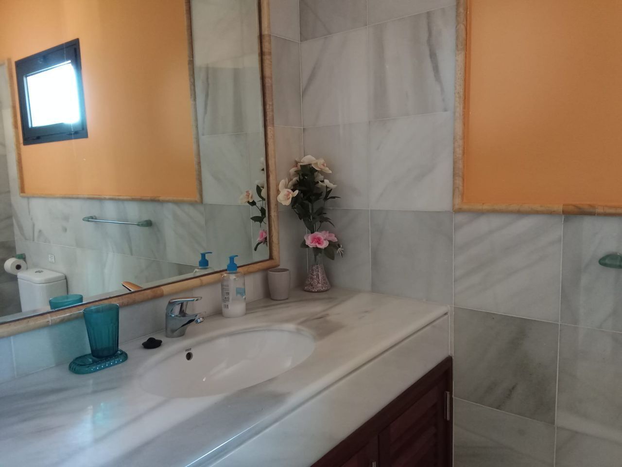 Een badkamer met een wit wastafel, een spiegel, een handdoekrek, een toilet en een vaas met bloemen.