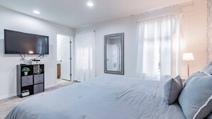 A bedroom with a large bed, a TV mounted on the wall, a bedside table with a lamp, and a door leading to another room.