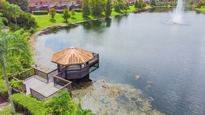 A lakeside gazebo with a wooden deck, surrounded by lush greenery and a calm lake.