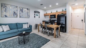 A modern living room with a blue sofa, a dining table with chairs, and a kitchen area with wooden cabinets and appliances.
