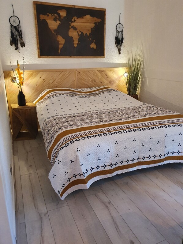 A neatly made bed with a patterned bedspread, a wooden headboard, and a world map above it. There are two wall-mounted dreamcatchers and a small plant beside the bed.