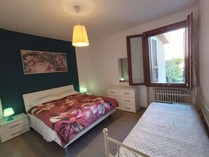 Ein Schlafzimmer mit Bett, Nachttischen, einem Schminktisch, einem Fenster mit Jalousien, einer Heizung und einer Pendelleuchte.