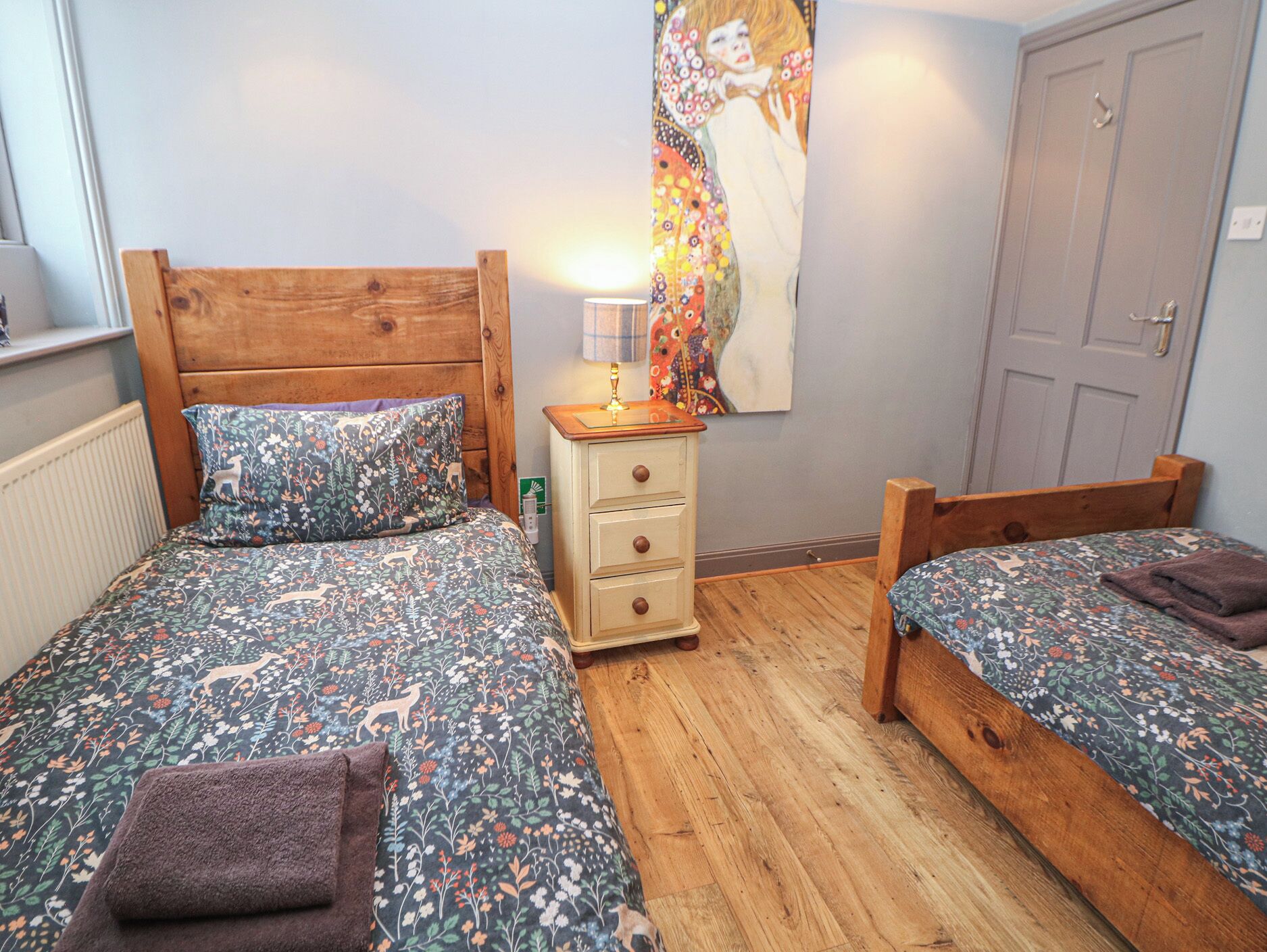 A bedroom with two single beds, a wooden headboard, a nightstand with a lamp, and a floral tapestry on the wall.