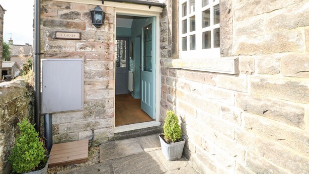 A stone building with a small entrance, a sign reading "9 LADIES", and a potted plant outside. - Nine Ladies (Matlock)