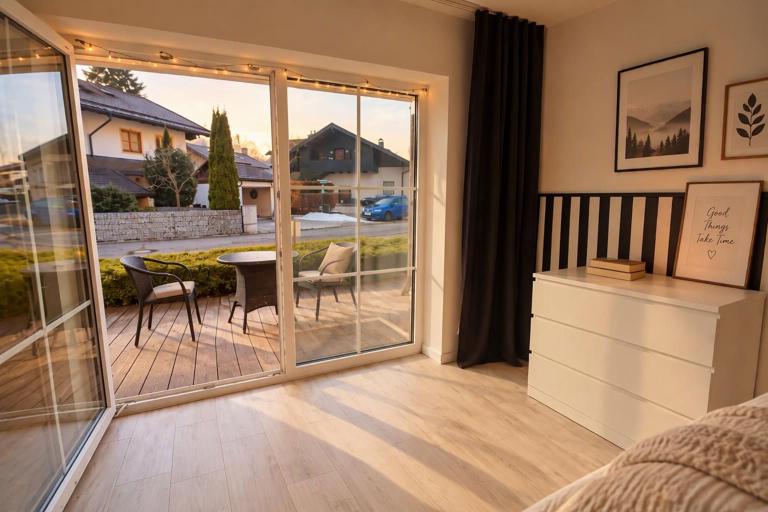 A modern bedroom with a sliding glass door leading to a balcony, a white dresser, and two framed pictures on the wall.