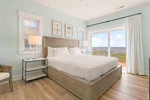 A bedroom with a large bed, a nightstand, a chair, and a view of the ocean through large windows. - Pier Envy 7 Bedroom home at Soundfront at Corolla Bay (Corolla)