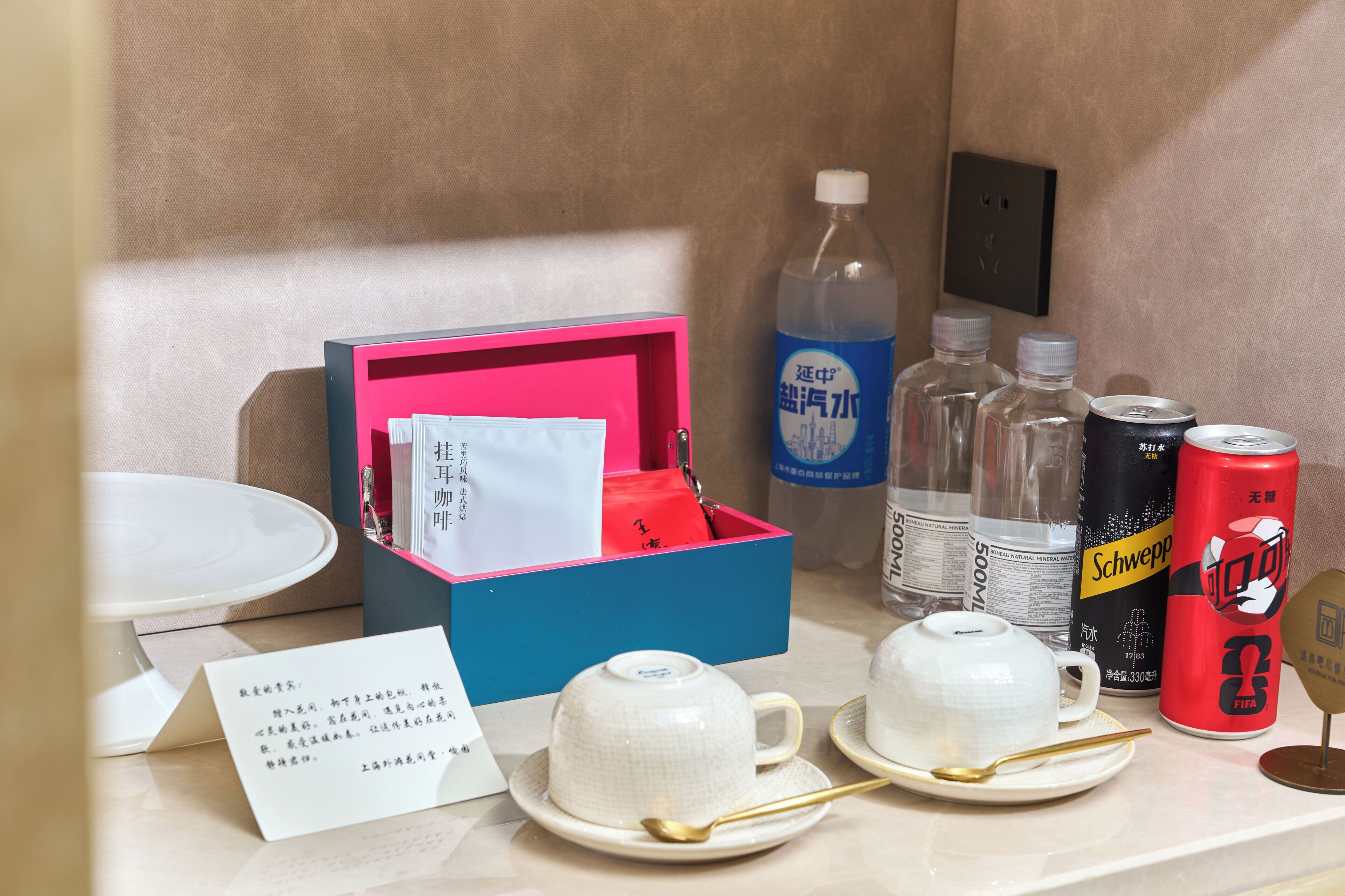 Une chambre d’hôtel avec une table mise, comprenant des tasses à thé, une cuillère, une boîte de conserve rouge, des bouteilles d’eau et une boîte rose contenant des mouchoirs.