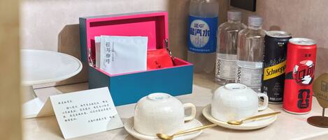 Habitación de hotel con una mesa dispuesta con tazas de té, una cuchara, una lata roja, botellas de agua y una caja rosa con pañuelos.