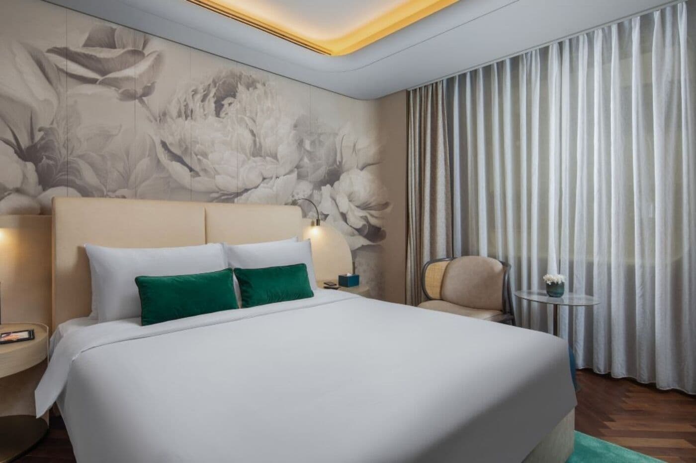 A hotel room with a large bed, a headboard, a wall mural, a chair, a small table, and a curtain.
