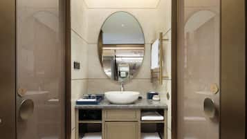 A modern bathroom with a large oval mirror, a white sink, and a marble countertop. There are two towel racks and a small shelf with toiletries.