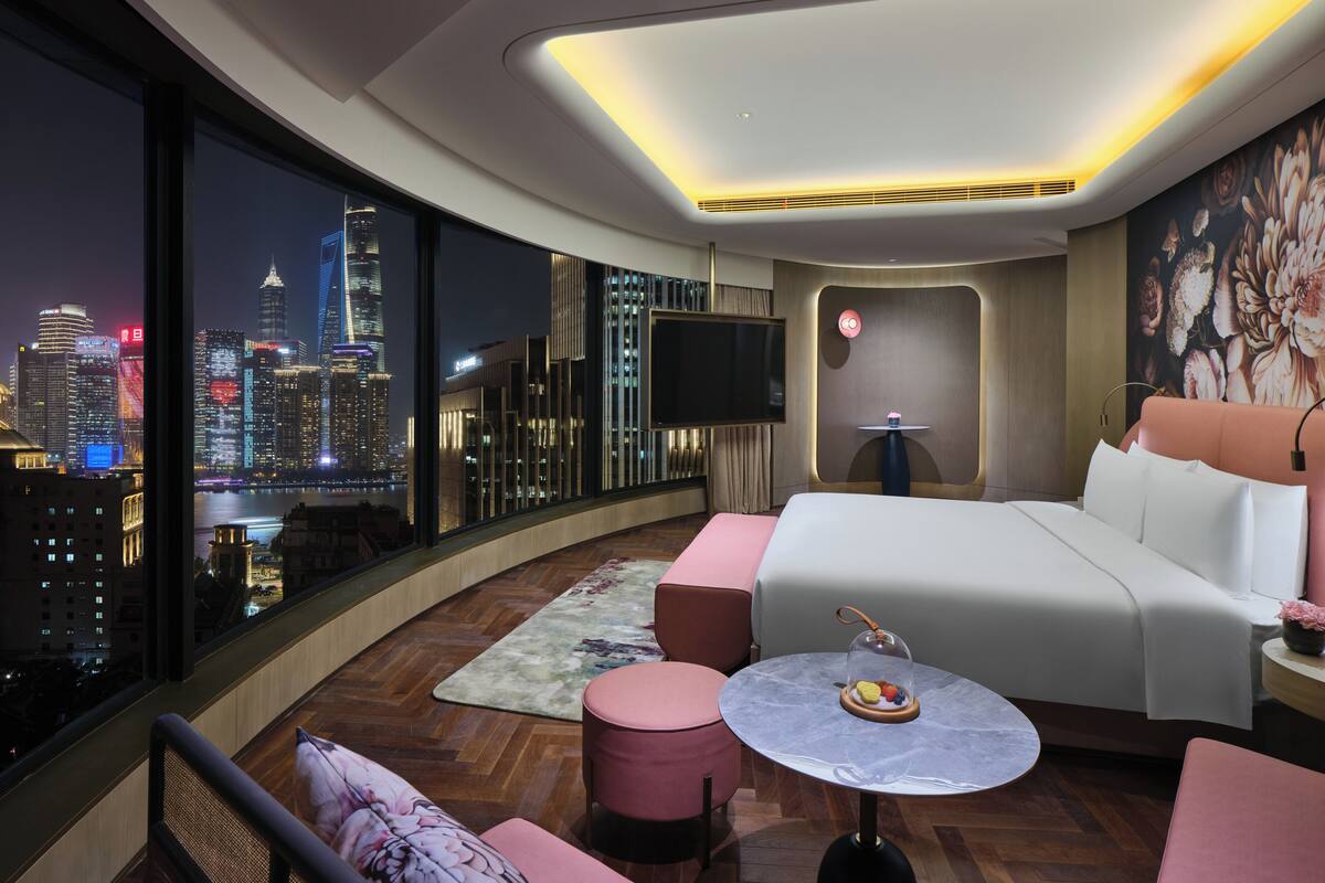Ein modernes Hotelzimmer mit einem großen Bett, einem runden Tisch mit Glaskuppel, einer Couch und Blick auf eine beleuchtete Stadtlandschaft bei Nacht.