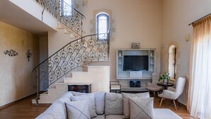 A spacious living room with a large beige sectional sofa, a staircase with ornate metal railings, a flat-screen TV mounted on the wall, and a small round table with chairs.