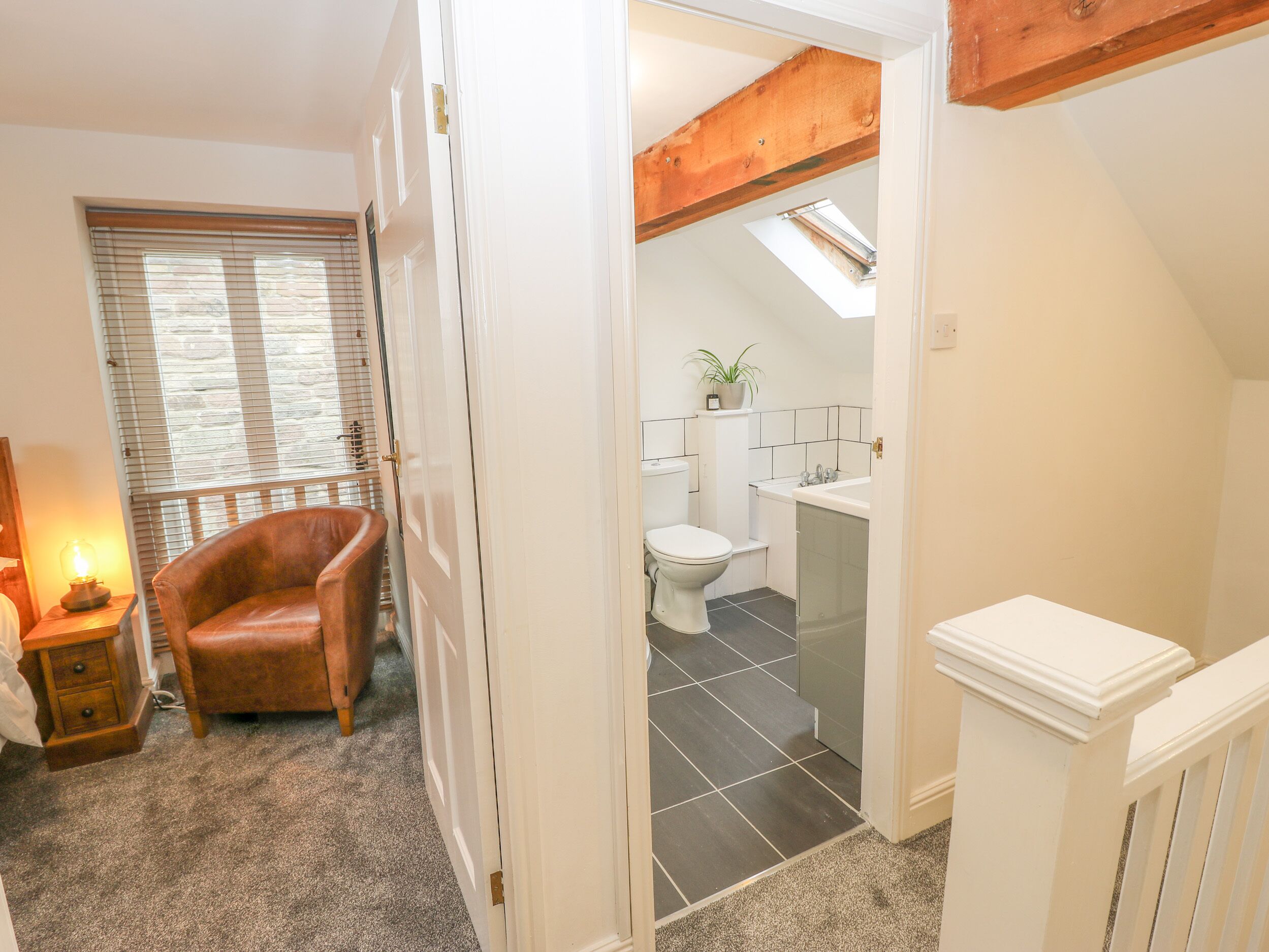 A hallway leading to a bathroom with a toilet, sink, and shower. There is a chair and a lamp in the hallway.