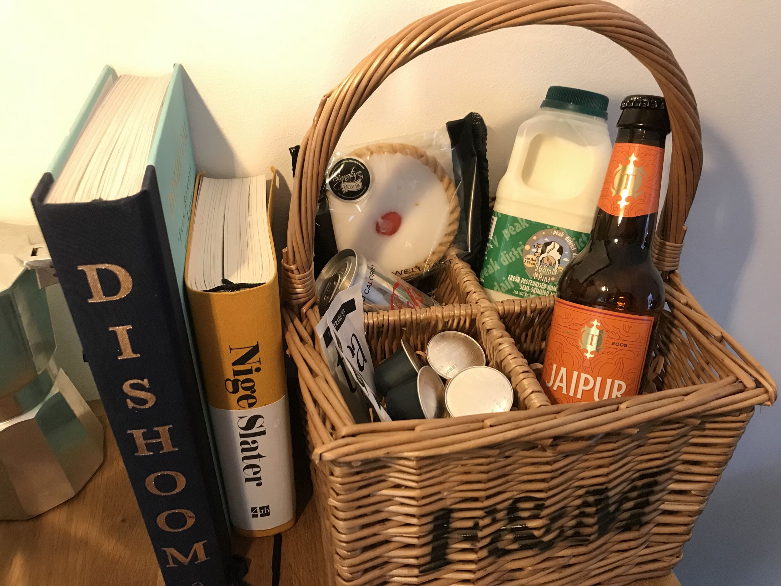 Un panier en osier contenant un livre intitulé « Dishome », une bouteille de bière « Jaipur », un emballage de lait et divers autres objets.