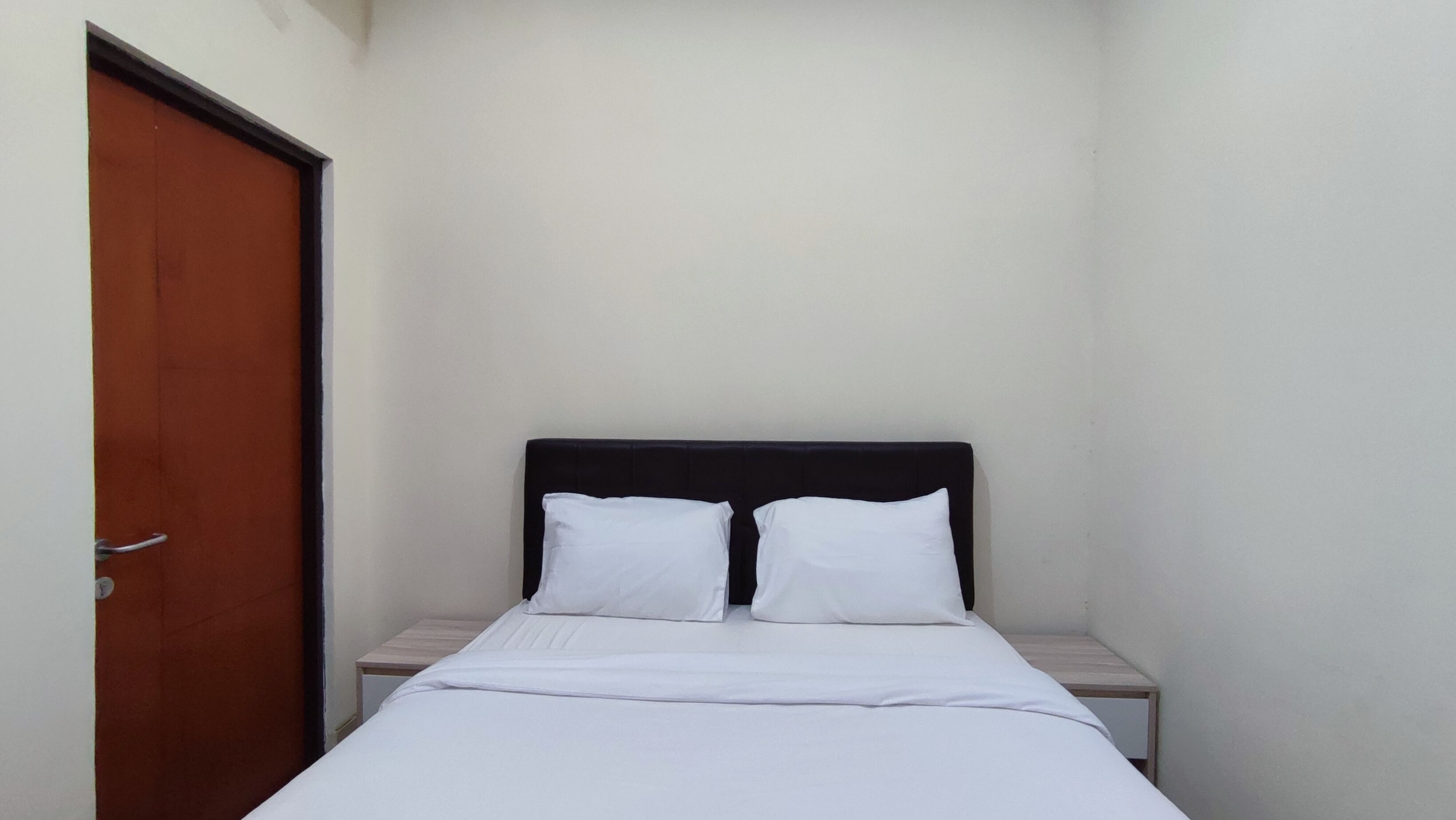 Una cama individual con ropa blanca en una habitación pequeña, con una puerta de madera y una mesita de noche.