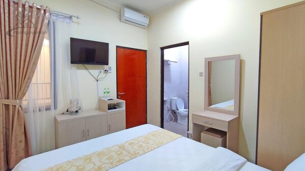 Una habitación de hotel con una cama, un televisor montado en la pared, una mesita con una botella, un espejo y una puerta que da a un baño.