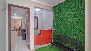 A reception area with a green wall, a wooden reception desk, and a staircase in the background. - Atmosfer Guest House Sentul by Ruang Nyaman (Sentul City)
