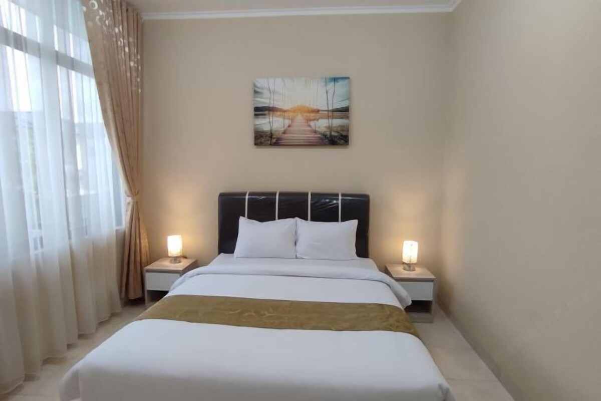 A hotel room with a large bed, two bedside tables with lamps, a painting on the wall, and sheer curtains.