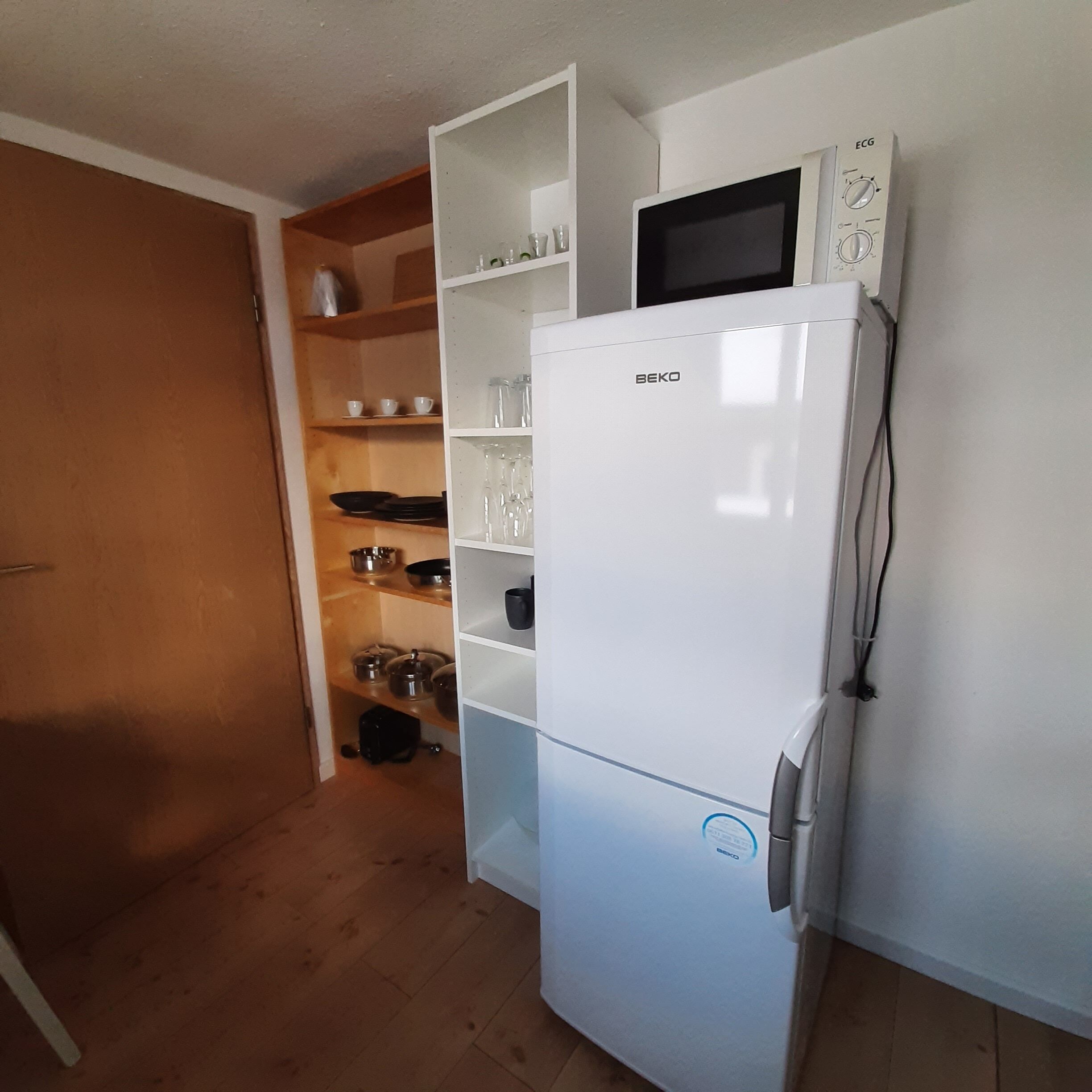 A compact kitchen with a white refrigerator, microwave, and a shelf unit with dishes and cookware.