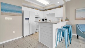 A modern kitchen with white cabinets, a built-in oven, and a refrigerator. There are blue bar stools and a painting of a beach on the wall. - Gulf 513-2 BED/2 BATH-sleeps 6-VIEWS OF POOL AND BEACH!  Updated (Fort Walton Beach)