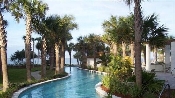 A curved swimming pool surrounded by palm trees and greenery, with a view of the ocean in the background. - Gulf 513-2 BED/2 BATH-sleeps 6-VIEWS OF POOL AND BEACH!  Updated (Fort Walton Beach)