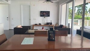 Una habitació àmplia amb una taula de dinar de fusta, cadires de vimetla, un sofà i un televisor fixat a la paret.