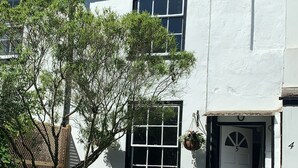 A white house with a chimney, a window, and a door. There is a tree in front of the house.
