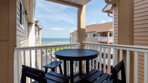Un balcon avec une table ronde et quatre chaises, donnant sur une plage et d’autres bâtiments.