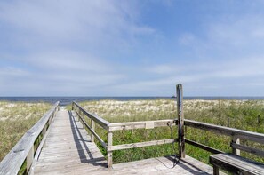 A wooden boardwalk leading to the beach with a bench and a sign on the right.