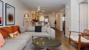 Un salon avec un canapé, une table basse en verre et un fauteuil. Au fond, on aperçoit une cuisine avec des placards et un réfrigérateur.