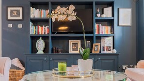 Un salon avec une table ronde en verre, un pouf capitonné, une bibliothèque et un téléviseur.