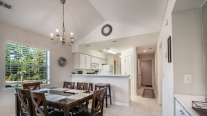 Un comedor con candelabro, una ventana con persianas, una cocina con armarios blancos y un pasillo que conduce a otra habitación.