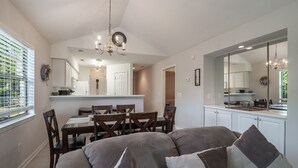 Una cocina moderna con armarios blancos, un comedor con mesa y sillas de madera, y un espejo grande que refleja la cocina.